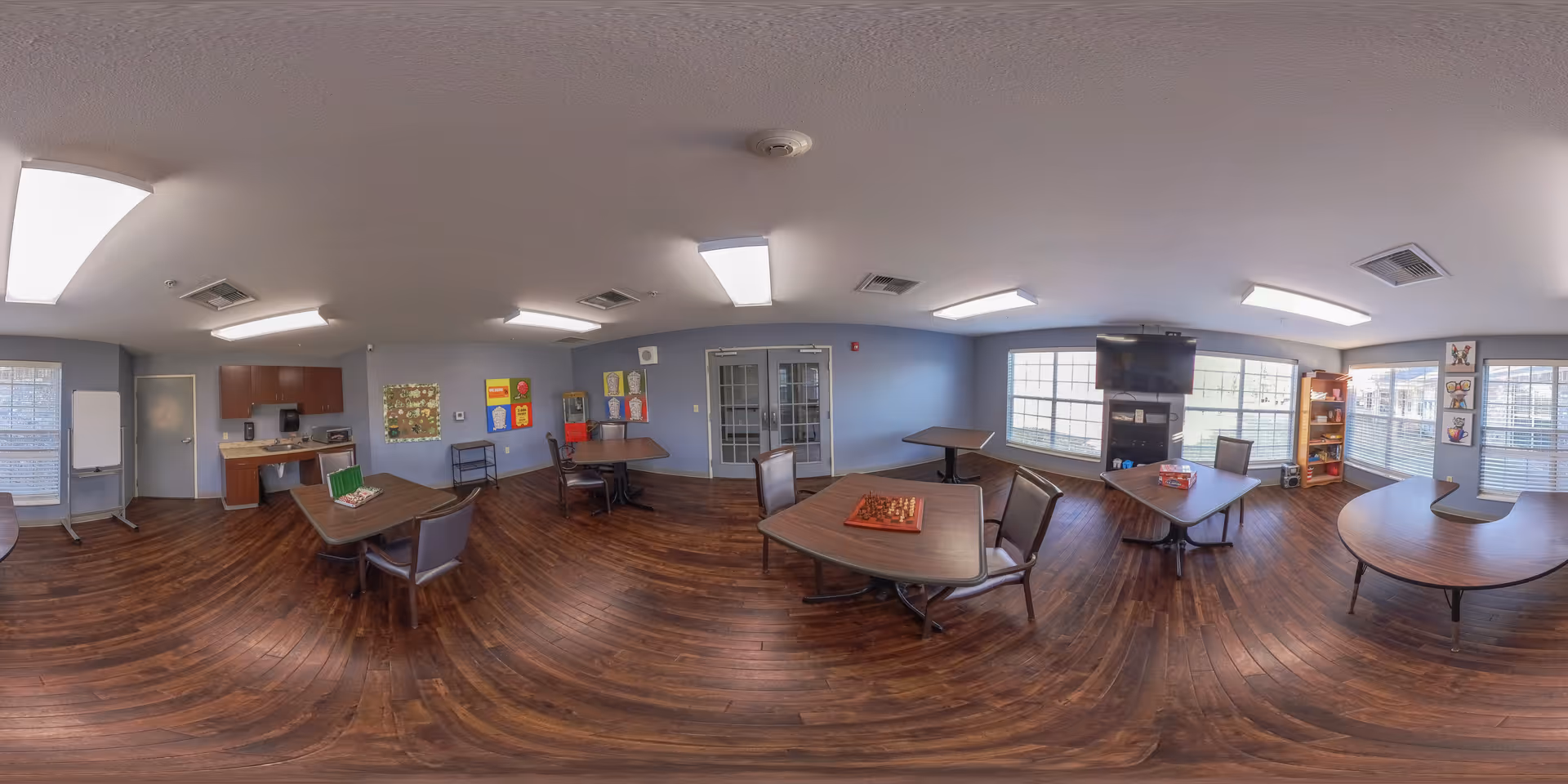 Spacious communal activity and dining room with multiple tables and chairs, a TV, kitchenette, and large windows.