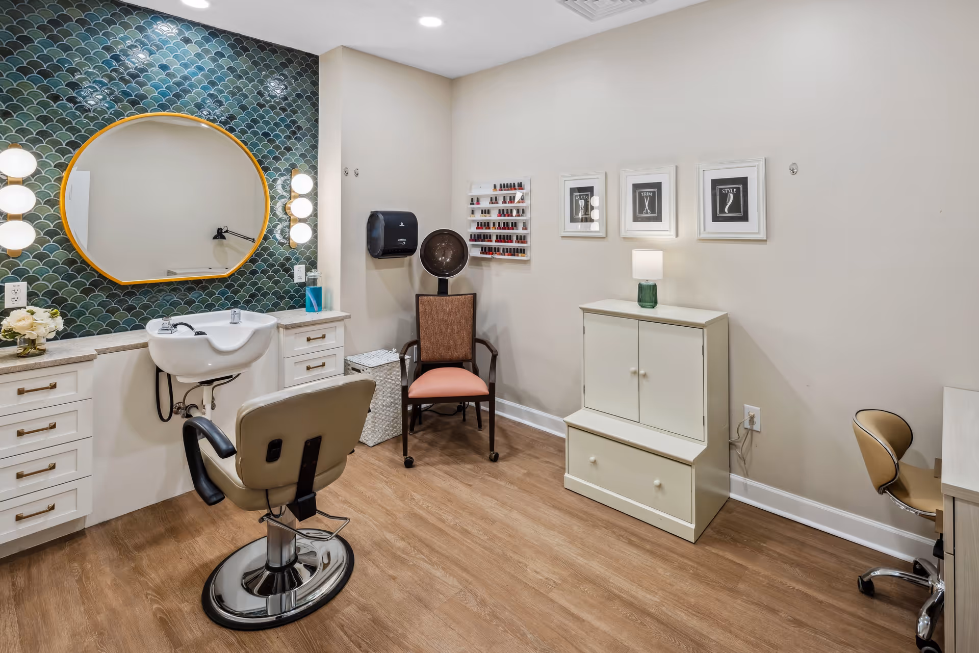 A beauty salon room with a styling chair in front of a counter with a sink and a large round mirror with lights on either side. The wall behind the mirror has green fish scale tiles. There is a hair dryer chair, a rack with nail polish bottles, a white cabinet with a small lamp on top, and three framed pictures on the wall. The floor is wood and the walls are light-colored.