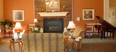 Cozy senior living lounge with brick fireplace, sofas, lamps, and a grand piano.