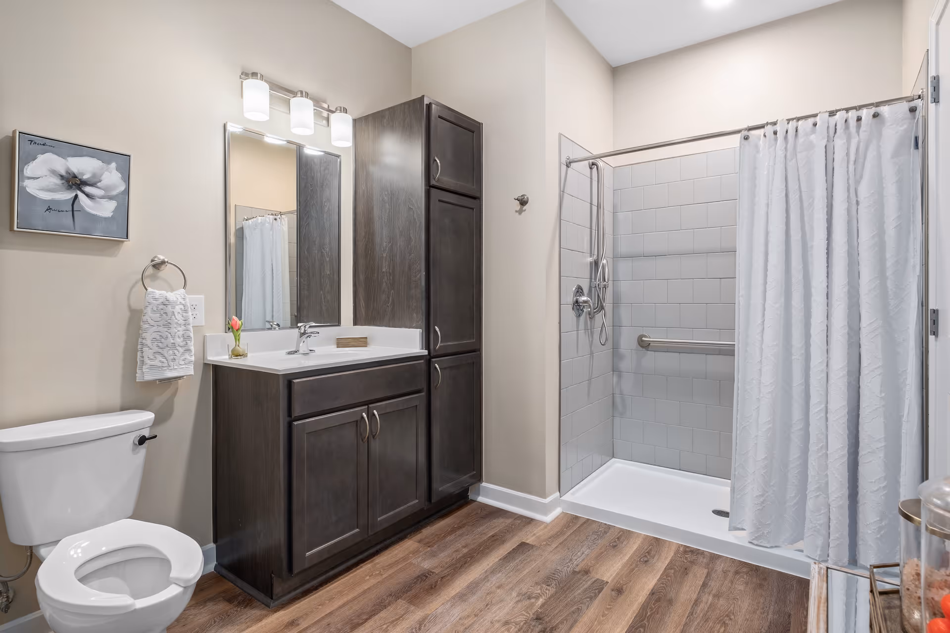 A clean and modern bathroom featuring a white toilet with an open seat, a dark wood vanity with a white countertop and a sink, a large mirror with three overhead lights, a tall matching cabinet, a walk-in shower with white tiles and a white shower curtain, and a small framed flower artwork on the wall.