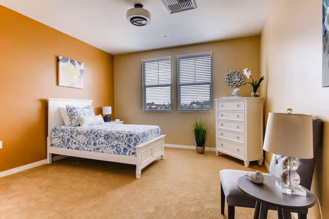 A bright bedroom with beige walls and carpet, featuring a white bed with blue and white patterned bedding, a white dresser with decorative items on top, a small round table with a lamp and chair, and two windows with blinds letting in natural light.