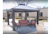 A covered outdoor gazebo with cushioned benches, chairs, and a table in a courtyard beside a single-story building.