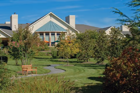 A senior living community building with a green lawn, trees, and a wooden bench along a winding pathway under a clear blue sky.