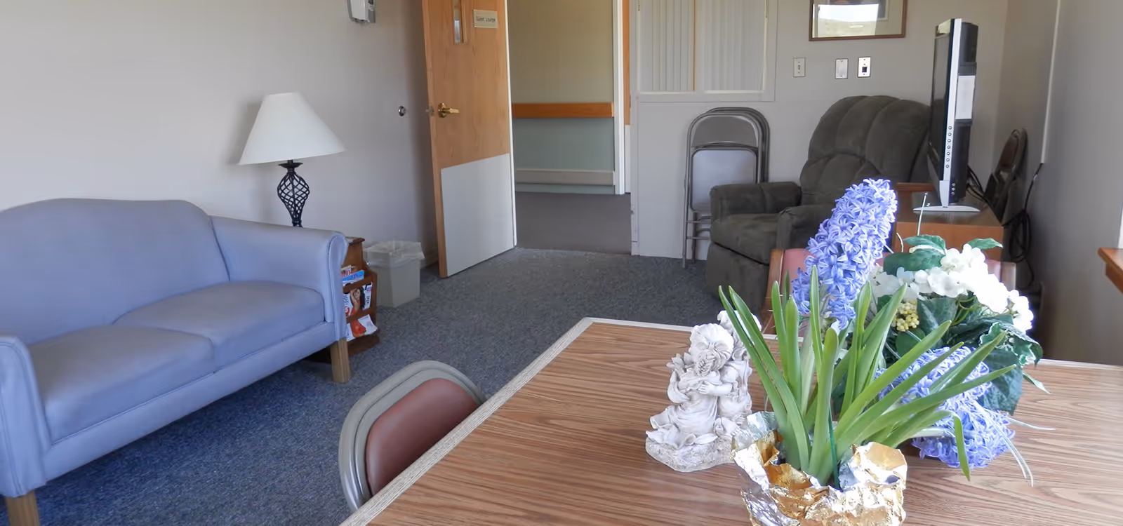 A cozy lounge area with a light blue couch, a table with a flower arrangement and a small angel statue, a green recliner chair, a TV on a stand, and a lamp on a side table. The door in the background has a sign that reads 'Guest Lounge'.