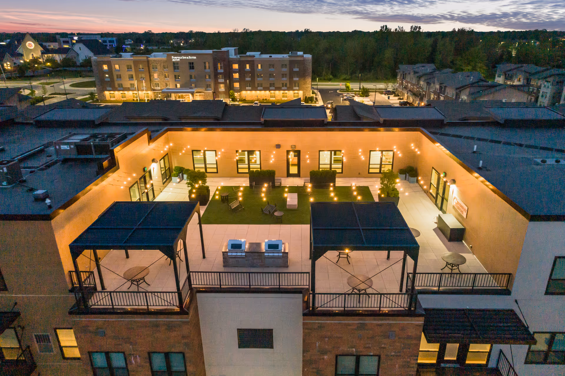 A rooftop courtyard at dusk with string lights, seating areas, pergolas, and surrounding buildings.