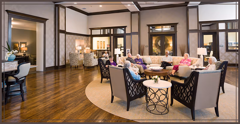 A group of elderly people sitting and socializing in a spacious, well-lit living room with wooden floors, beige walls, and comfortable armchairs and sofas arranged around a circular rug and coffee table.