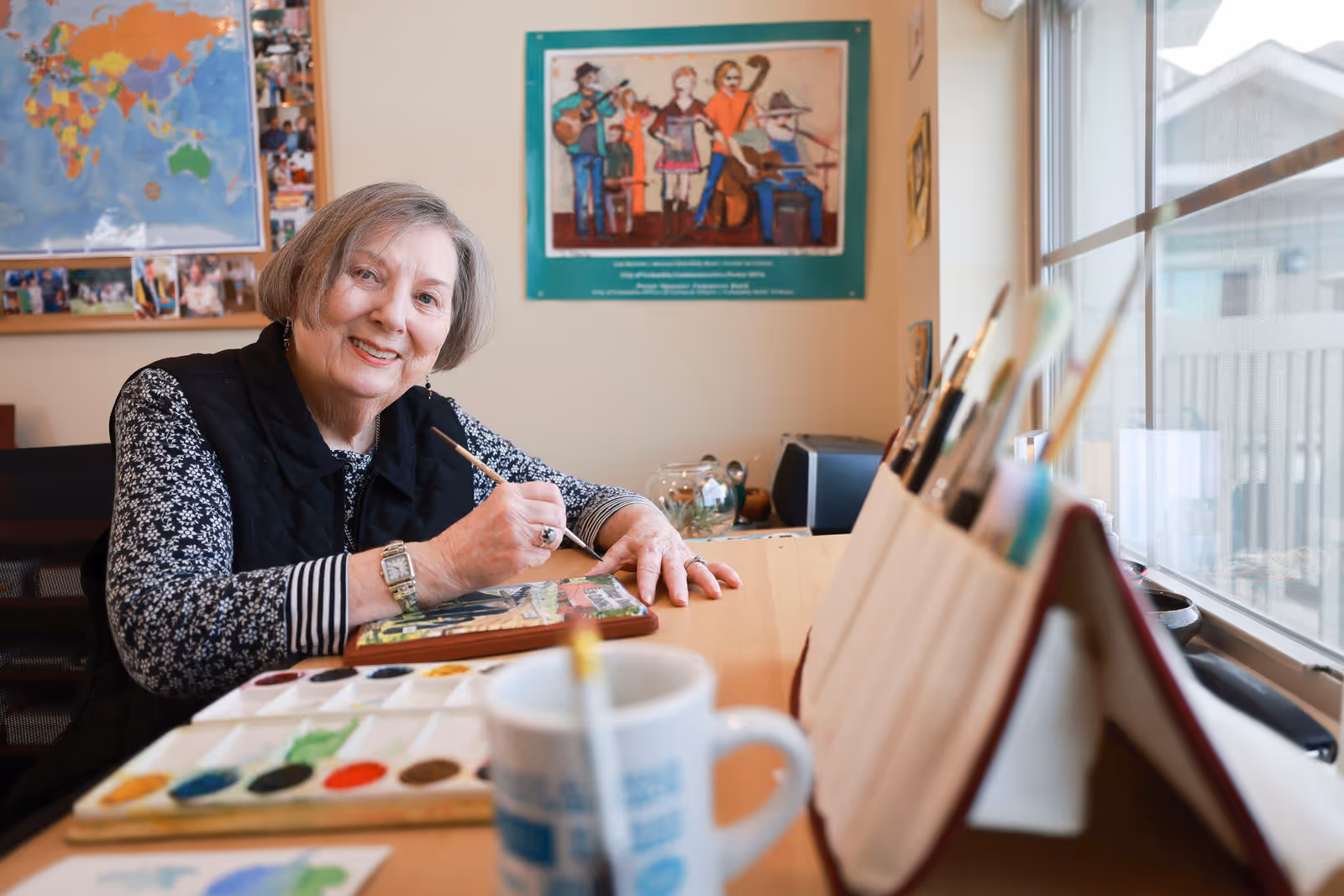 An elderly woman sitting at a table by a window, smiling while painting with watercolors. Various paintbrushes are in a holder on the table, and a palette with multiple colors is in front of her. Behind her on the wall are a world map and a colorful poster featuring musicians.