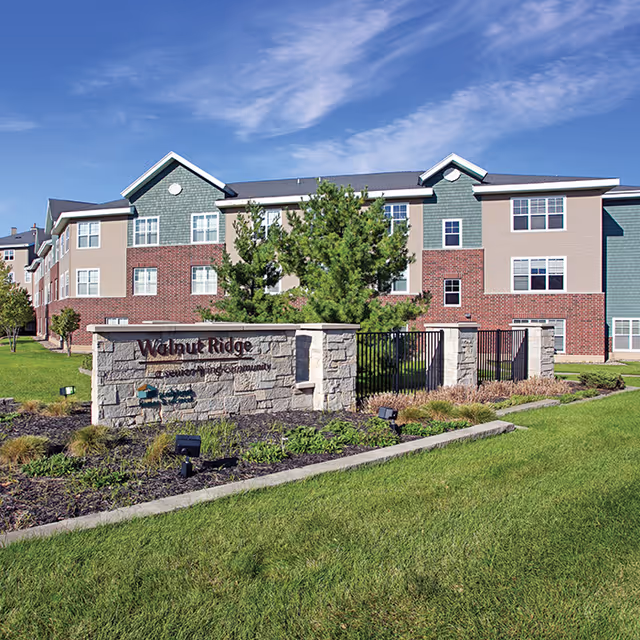 Exterior view of Walnut Ridge senior living community building with a stone sign in front displaying the facility name. The building has multiple windows, a combination of brick and siding exterior, and is surrounded by green grass and landscaping under a blue sky with some clouds.