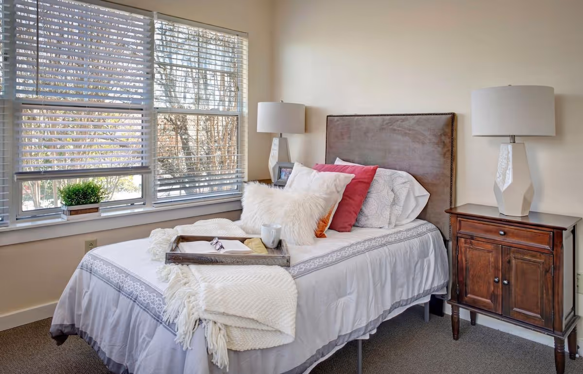 A neatly made bed with white and pink pillows, a white throw blanket, and a wooden tray holding a book, glasses, and a mug. The bed is positioned next to a large window with white blinds, and there is a wooden nightstand with a white lamp on it beside the bed.