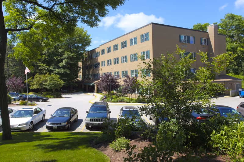 Exterior view of a multi-story brick building with multiple windows, surrounded by trees and greenery. Several cars are parked in the parking lot in front of the building under a clear blue sky.