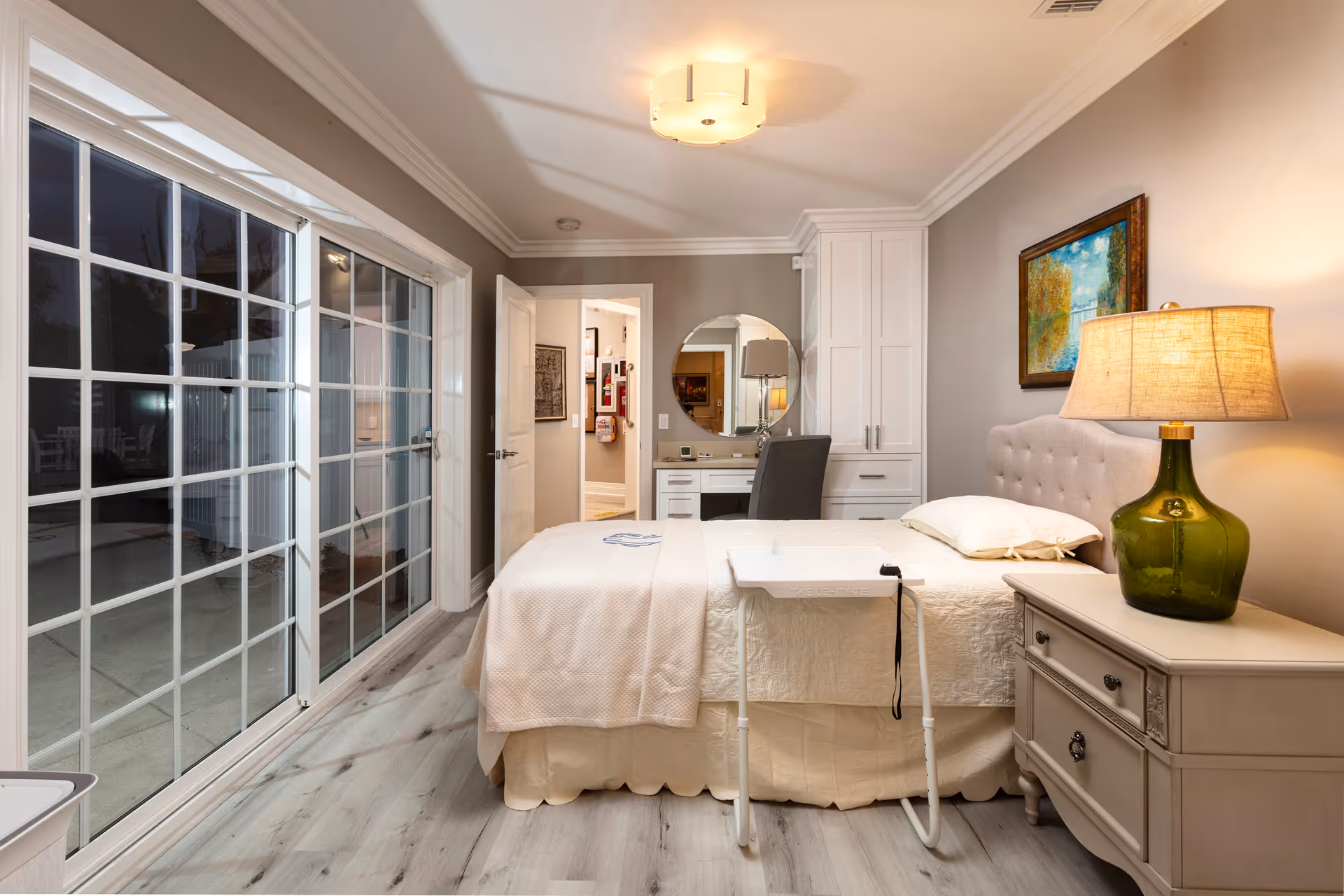 A cozy bedroom with a bed covered in cream-colored bedding and a small white overbed table. To the right of the bed is a nightstand with a green glass lamp and a framed painting on the wall above the bed's tufted headboard. At the far end of the room is a white desk with a round mirror and a gray chair. Large sliding glass doors on the left side open to an outdoor patio area. The room has light wood flooring and soft gray walls with white trim.