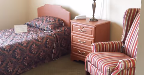 A small bedroom with a single bed covered in a patterned bedspread, a wooden nightstand with three drawers, a lamp, and a telephone on top. Next to the nightstand is a striped upholstered armchair.