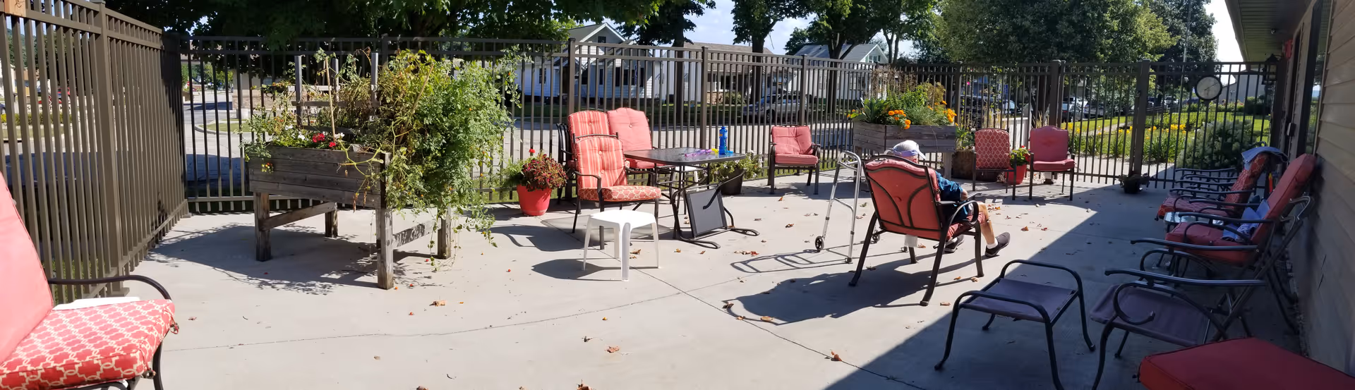 Outdoor patio area with several cushioned chairs and small tables arranged on a concrete surface. There is a raised garden bed with plants, a metal fence enclosing the area, and trees providing shade. Two elderly individuals are seated in chairs, enjoying the outdoor space.