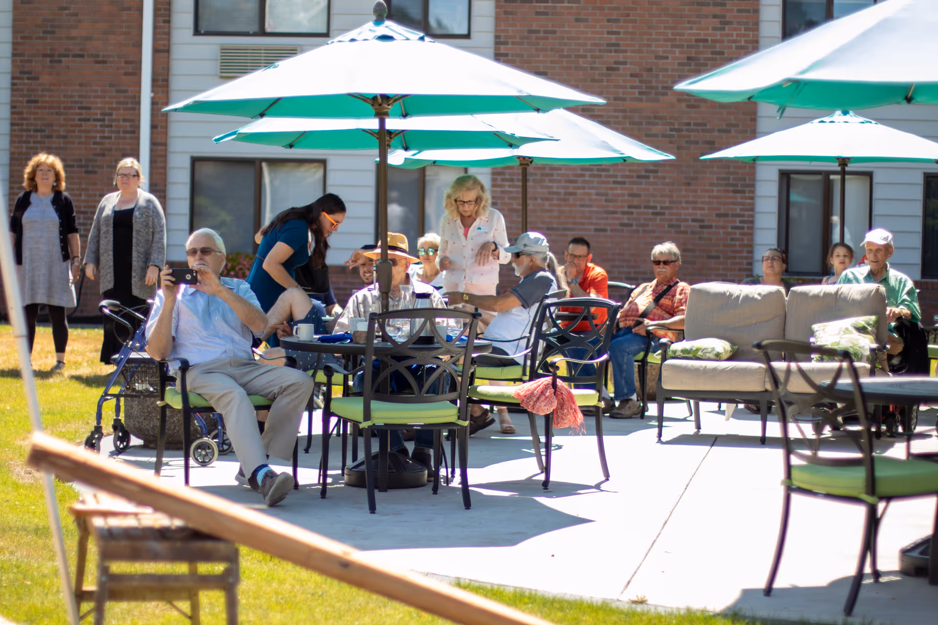A group of elderly people sitting and standing outdoors on a sunny day at King City Senior Village. They are gathered around tables with green umbrellas and some are seated on outdoor chairs and sofas on a concrete patio in front of a brick building.