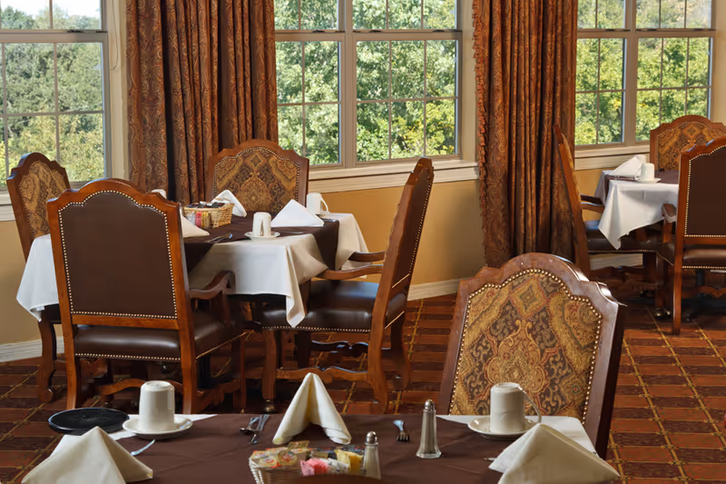 Dining area with tables covered in white and brown tablecloths, set with cups, napkins, and condiments. The room has large windows with brown patterned curtains and wooden chairs with upholstered backs and seats.