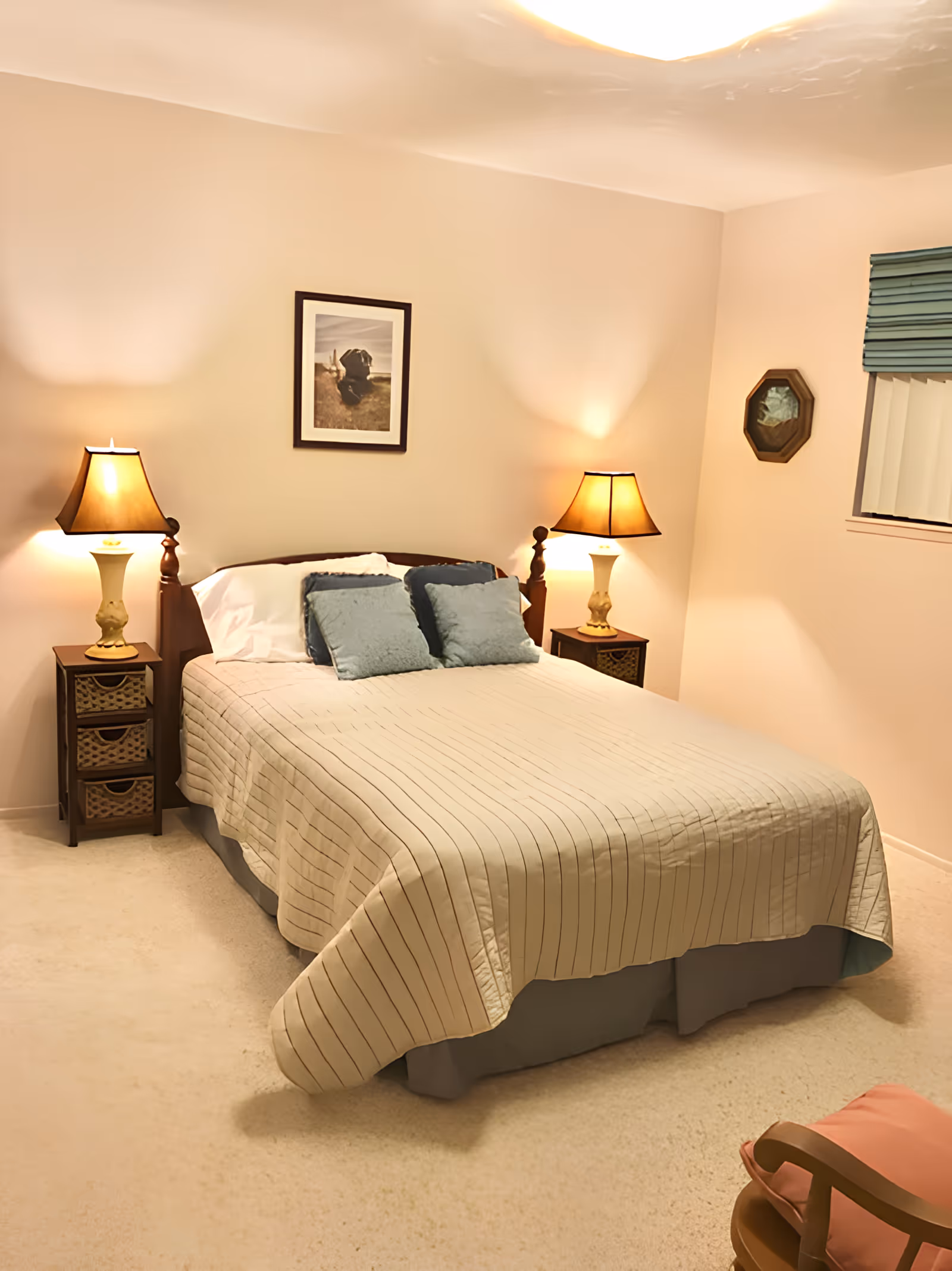 A cozy bedroom with a neatly made bed featuring a beige striped bedspread and three blue pillows. There are two wooden nightstands on either side of the bed, each with a lamp emitting warm light. A framed picture of a dog hangs on the wall above the bed. The room has light-colored walls and carpet, a window with blue blinds, and a wooden chair with a pink cushion in the corner.