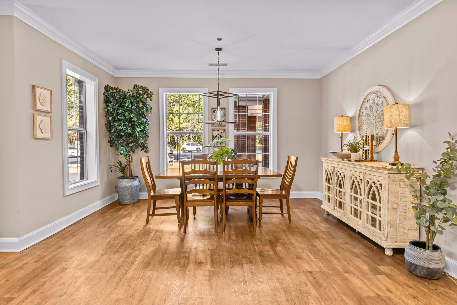 A bright dining room with a wooden table and six matching chairs. The room has light beige walls, two large windows letting in natural light, a potted plant in the corner, and a decorative sideboard with two lamps and a round wall decoration above it. The floor is wooden, and the overall atmosphere is warm and inviting.