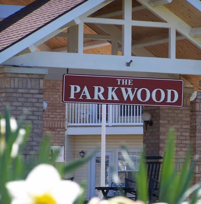 Entrance sign reading 'The Parkwood' hanging under a wooden roof structure with brick pillars, with blurred flowers in the foreground and part of a building with a balcony and door in the background.