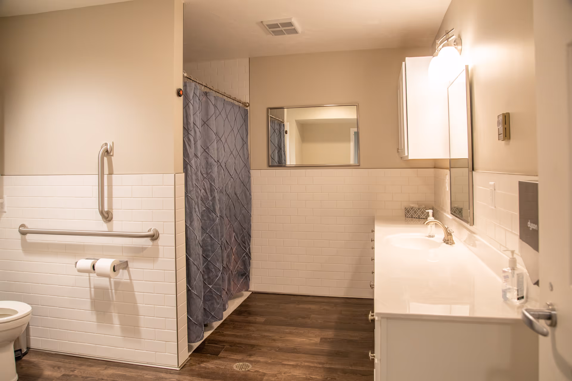 A clean and spacious senior care bathroom with white tiled walls, a toilet with grab bars, a shower with a gray curtain, a large mirror above a white sink countertop, and wooden flooring.