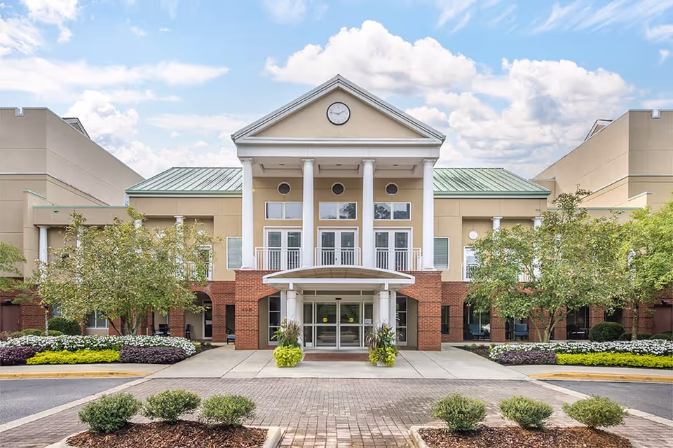 Front exterior view of a senior living facility building with a symmetrical design, featuring a central entrance with glass doors, white columns, a clock above the entrance, and landscaped greenery including trees and flower beds on either side.