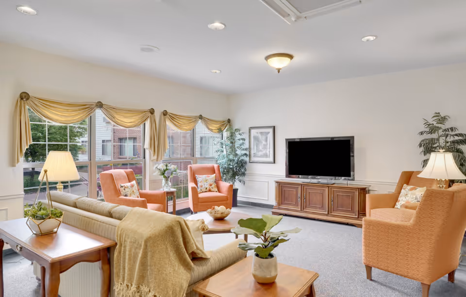 Bright communal living room with sofa, upholstered armchairs, coffee tables, TV cabinet and large windows.