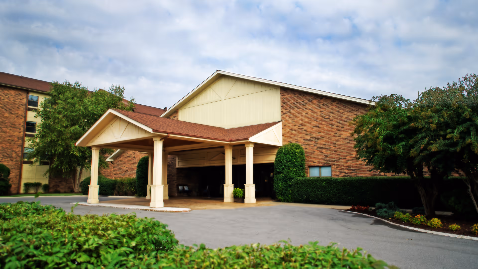 Covered porte-cochère entrance of a brick retirement community building with landscaping and a driveway.