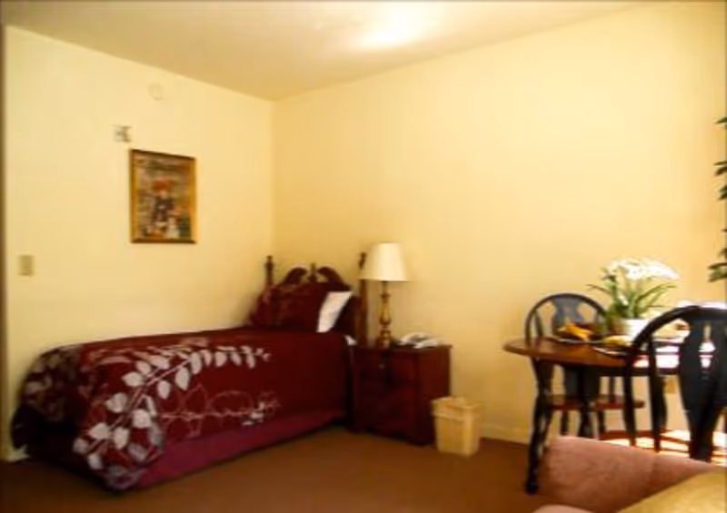 A small room with a single bed, bedside table and lamp, and a round dining table with chairs and a vase of flowers.