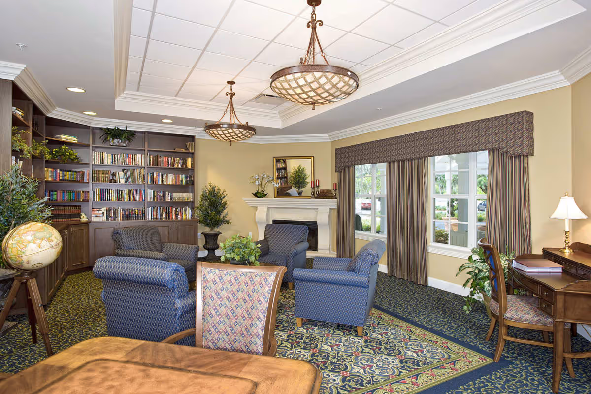 A cozy living room area in a senior living facility featuring four blue upholstered armchairs arranged around a small table with a plant centerpiece. The room has a patterned carpet, a fireplace with decorative items on the mantel, large windows with striped curtains, built-in bookshelves filled with books, and a wooden desk with a chair and lamp. Two ceiling light fixtures hang from a white tray ceiling.