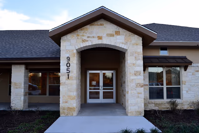 Front entrance of a building with stone pillars and an arched entryway. The building has beige stone and tan walls with a dark shingled roof. The number 9051 is displayed vertically on the left pillar. There are glass double doors at the entrance and windows on either side.