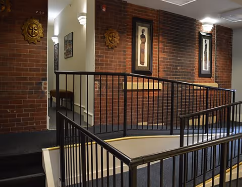 Interior view of a hallway with brick walls, black metal railings along a staircase, decorative wall art including framed pictures and sun-shaped wall sculptures, and carpeted flooring.