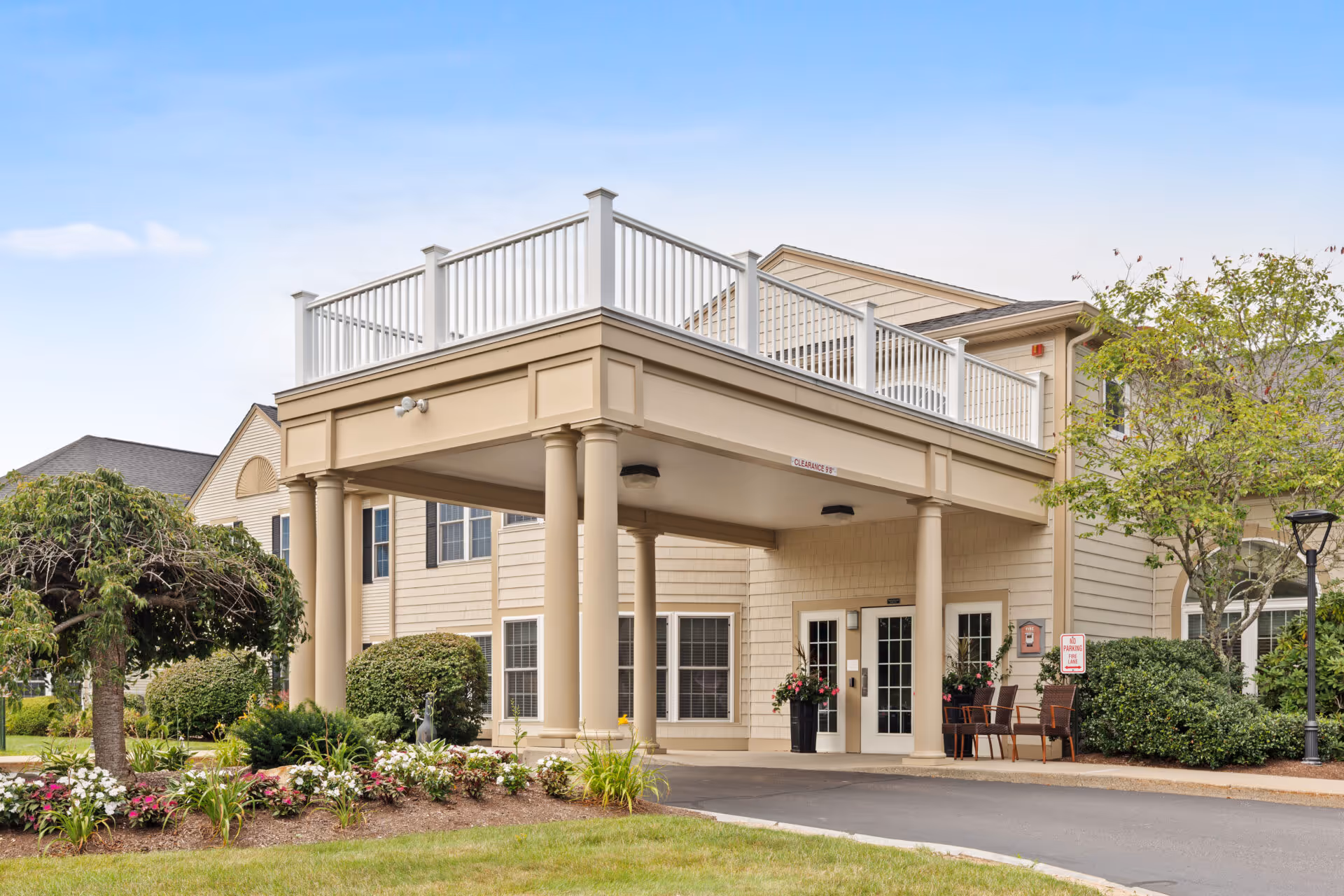 Exterior view of Benchmark Senior Living at Plymouth Crossings showing the entrance with a covered driveway supported by columns, surrounded by landscaped greenery and flowers under a clear blue sky.