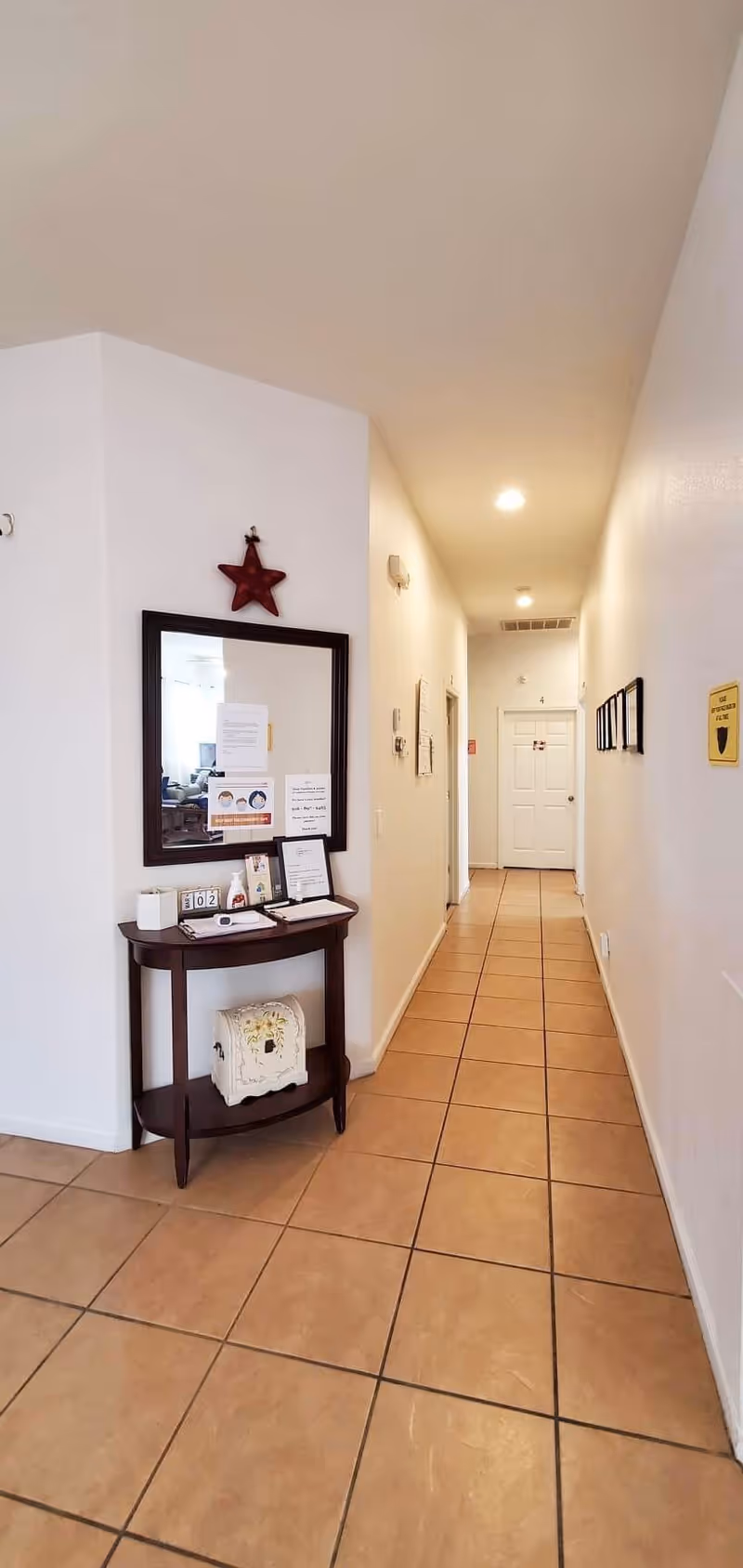 A long hallway with beige tiled floor and white walls, featuring a small dark wooden table with a mirror above it on the left side. The table holds various papers, a calendar, and a decorative item. The hallway is well-lit with ceiling lights and has several doors along the right side and at the end.