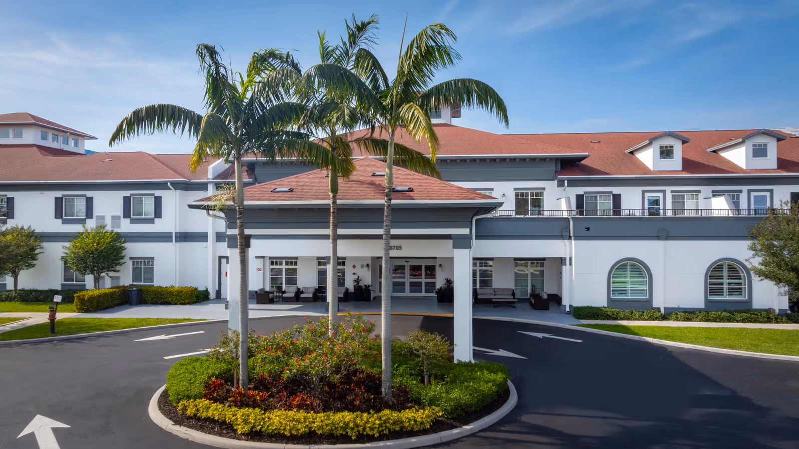 Front exterior of the Alamar Senior Living building with a covered entrance, palm trees, and a landscaped circular driveway.