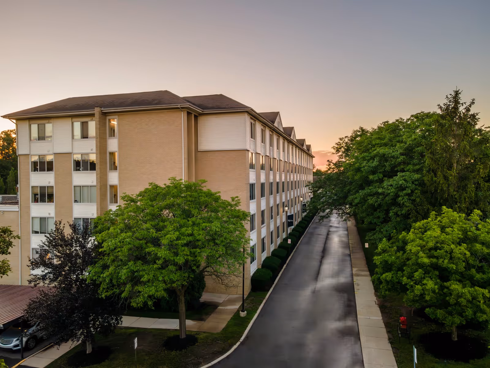 Exterior view of a multi-story senior living facility building at sunset with a driveway and lush green trees on both sides.