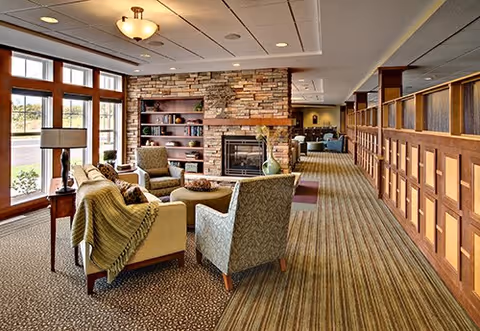 A cozy living room area in a senior living facility featuring a stone fireplace, built-in wooden shelves with decorative items, several upholstered chairs and a sofa arranged around a central coffee table, large windows letting in natural light, and a carpeted floor with patterned and striped designs.