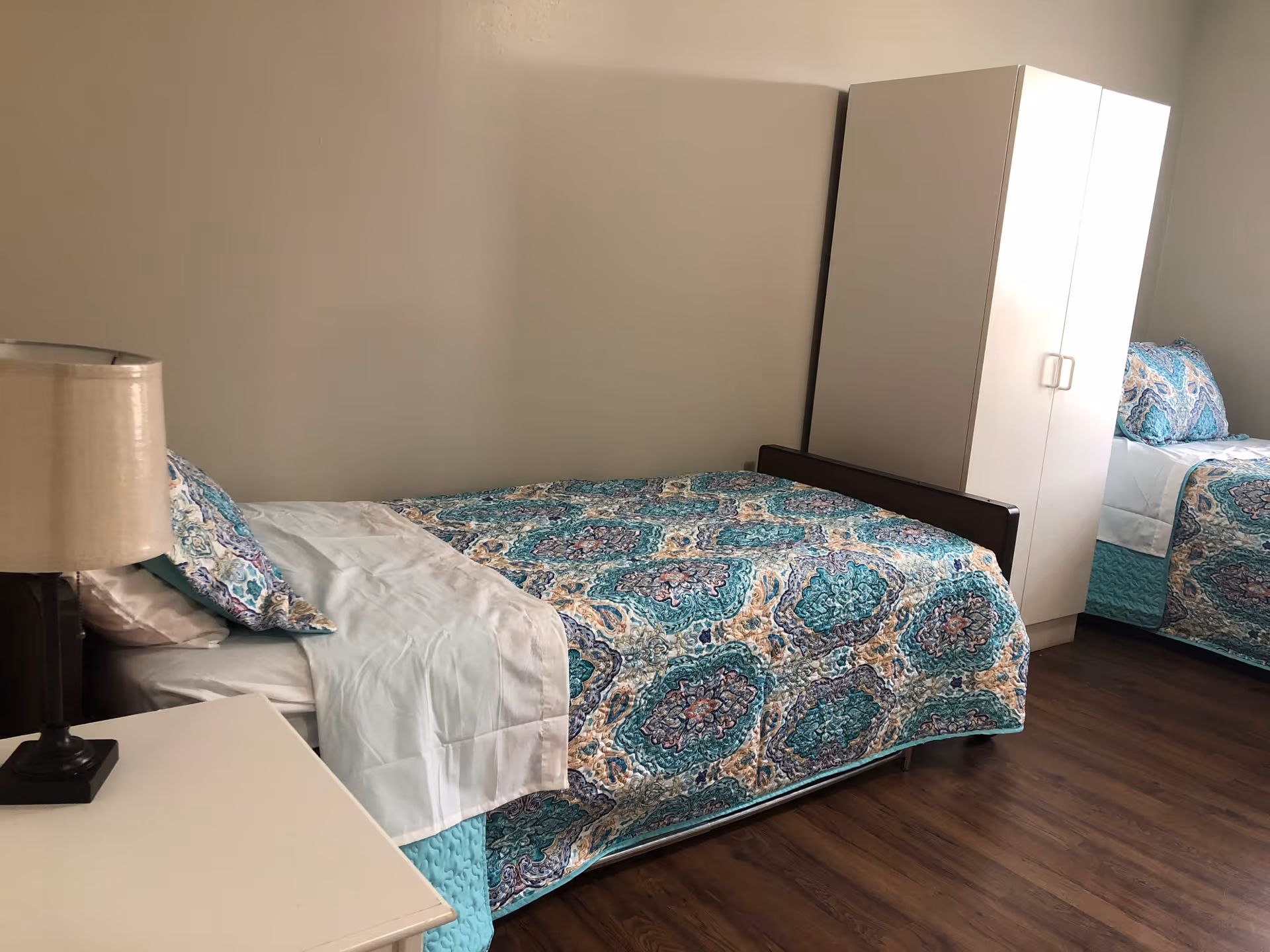 A bedroom with two single beds, each covered with a blue and teal patterned quilt. There is a white nightstand with a lamp on it next to one of the beds, and a white wardrobe is positioned between the beds. The room has wooden flooring and plain light-colored walls.