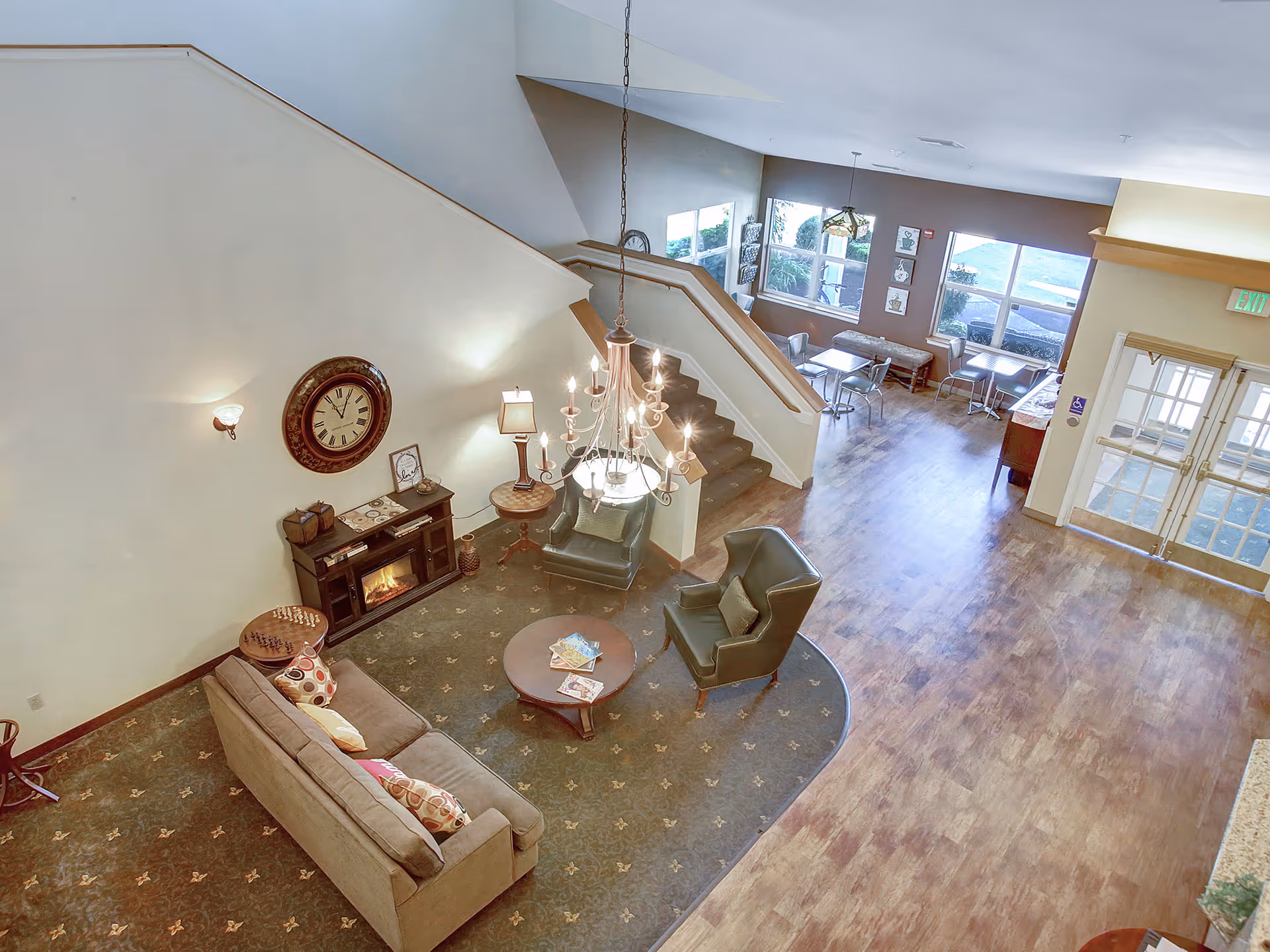 Spacious senior living lobby with a seating area around a coffee table and fireplace, a central chandelier, staircase, and dining tables by large windows.