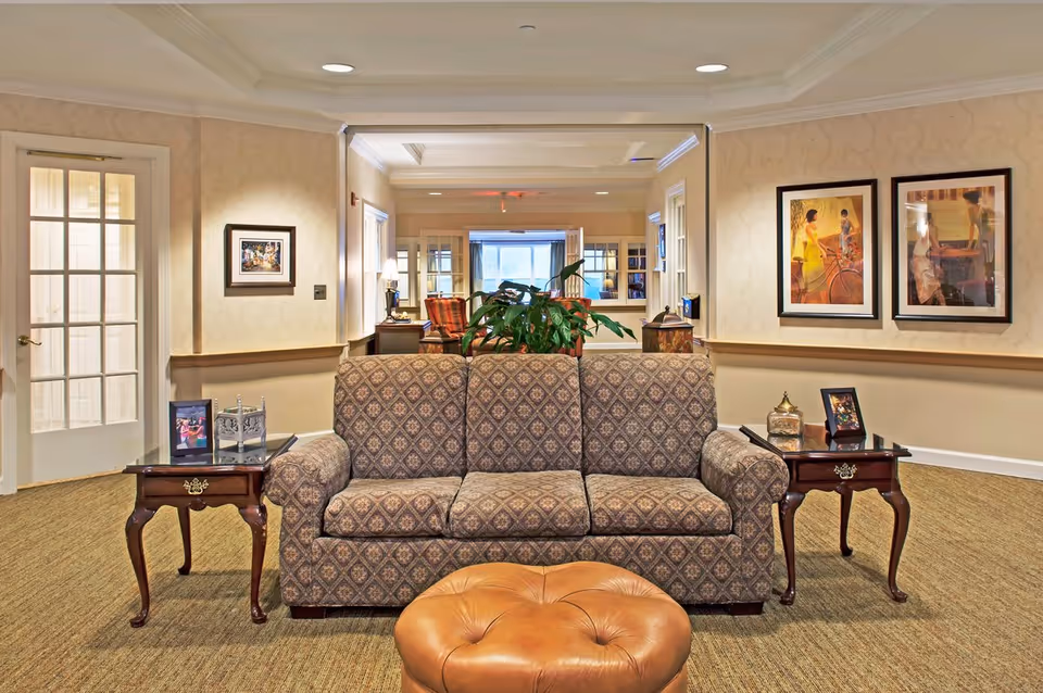 A cozy senior living facility common area with a patterned three-seater sofa centered between two wooden side tables. Each table holds decorative items including framed photos and a lantern. Behind the sofa is a large green plant, and the room features warm beige walls adorned with framed artwork. A glass-paneled door is visible on the left, and the space extends into another seating area with armchairs and a window in the background.