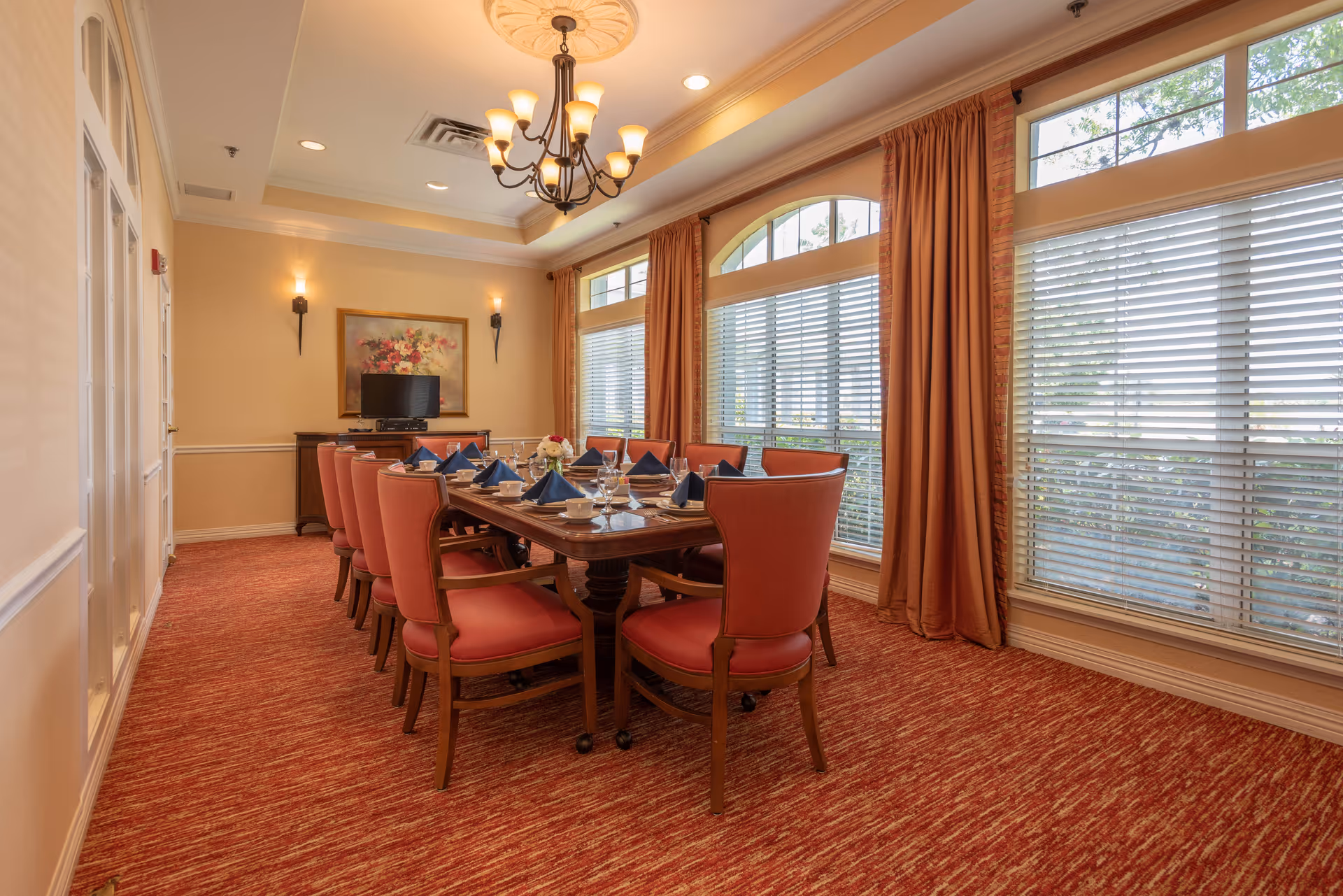 A formal dining room with a long wooden table set for a meal, surrounded by red upholstered chairs. The table is set with blue napkins, plates, glasses, and a floral centerpiece. Large windows with blinds and orange curtains allow natural light to fill the room. The walls are beige with white trim, and there is a chandelier hanging from the ceiling along with wall sconces. A sideboard with a TV and a floral painting is visible at the far end of the room.