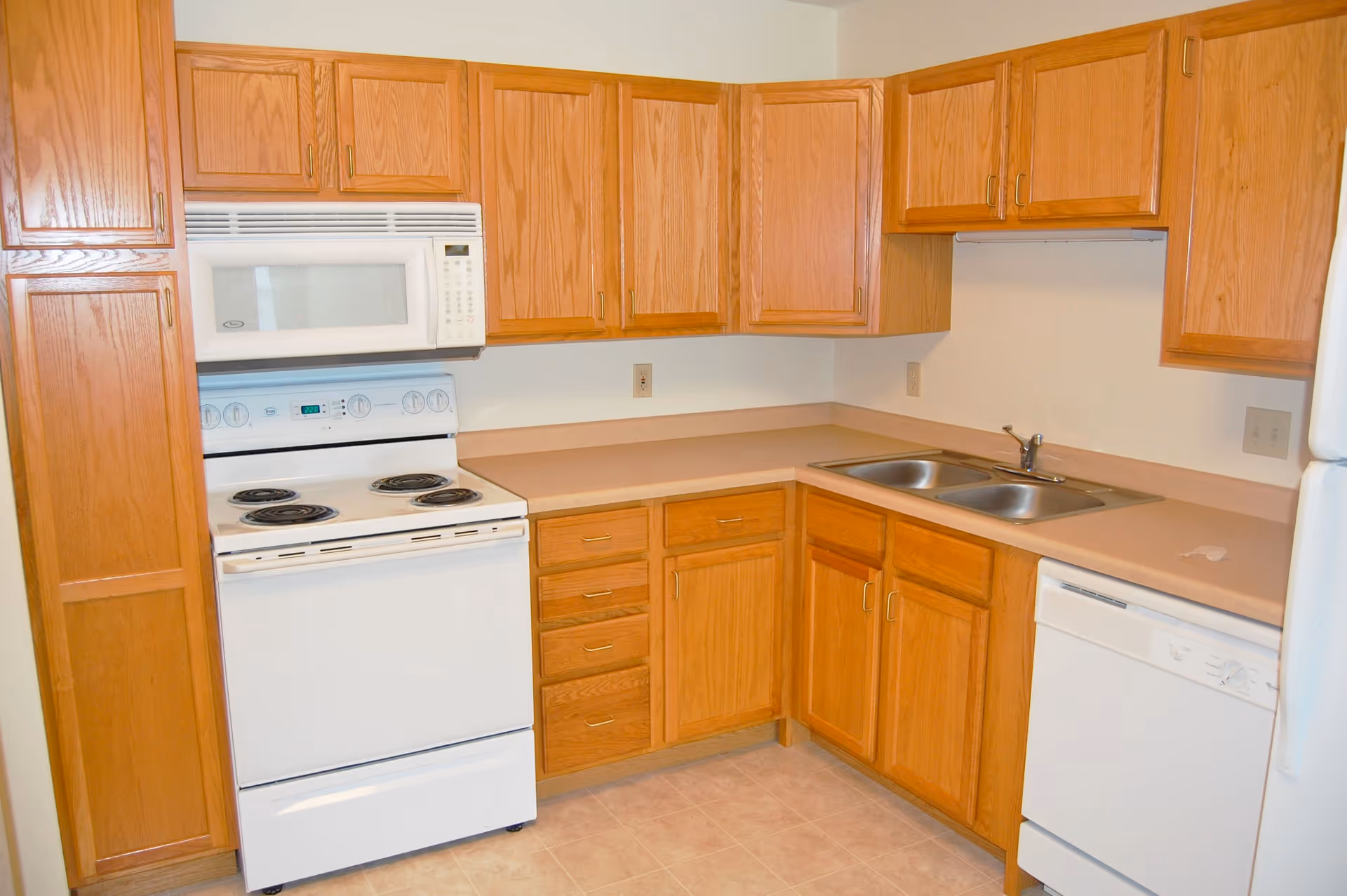 A kitchen with wooden cabinets, a white electric stove with four burners, a white microwave above the stove, a double stainless steel sink, a white dishwasher, and beige countertops and tiled floor.
