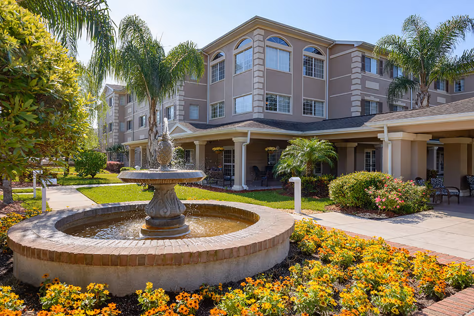 Courtyard of a senior living facility featuring a central fountain, colorful flower beds, palm trees, and the multi-story building entrance in the background.