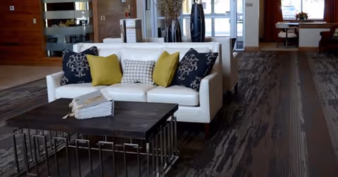 A modern living room area with a white sofa adorned with decorative pillows in black, yellow, and patterned designs. In front of the sofa is a dark wooden coffee table with a stack of books. The room features wood-paneled walls, large windows, and a carpeted floor with a striped pattern. In the background, there is a dining area with a table and chairs near the windows.
