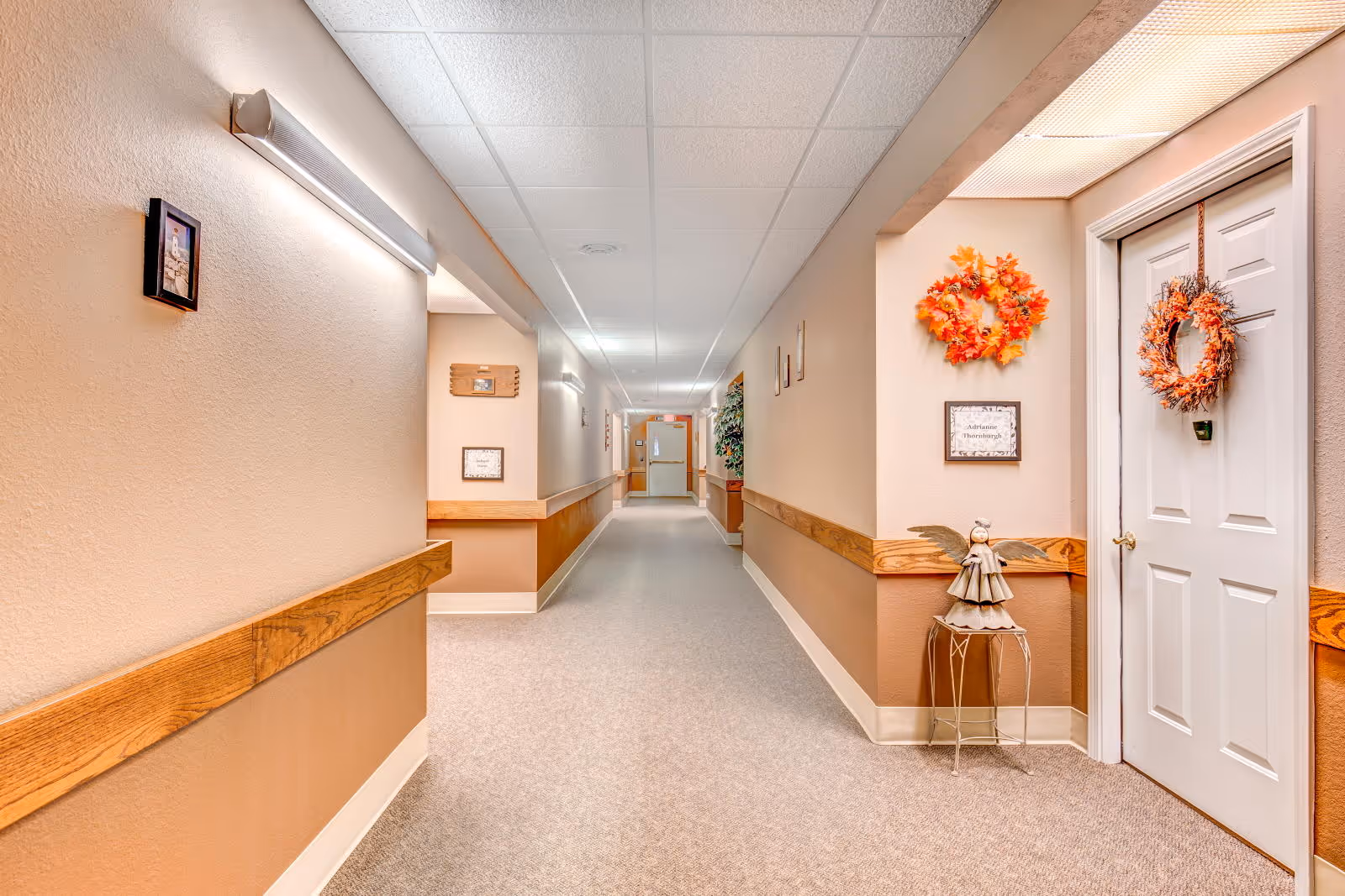 A well-lit hallway in a senior living facility with beige walls and carpeted floor. The hallway features wooden handrails on both sides, framed pictures on the walls, and decorative wreaths hanging on a white door to the right. A small angel figurine sits on a metal stand near the door. The ceiling has white tiles with fluorescent lighting.