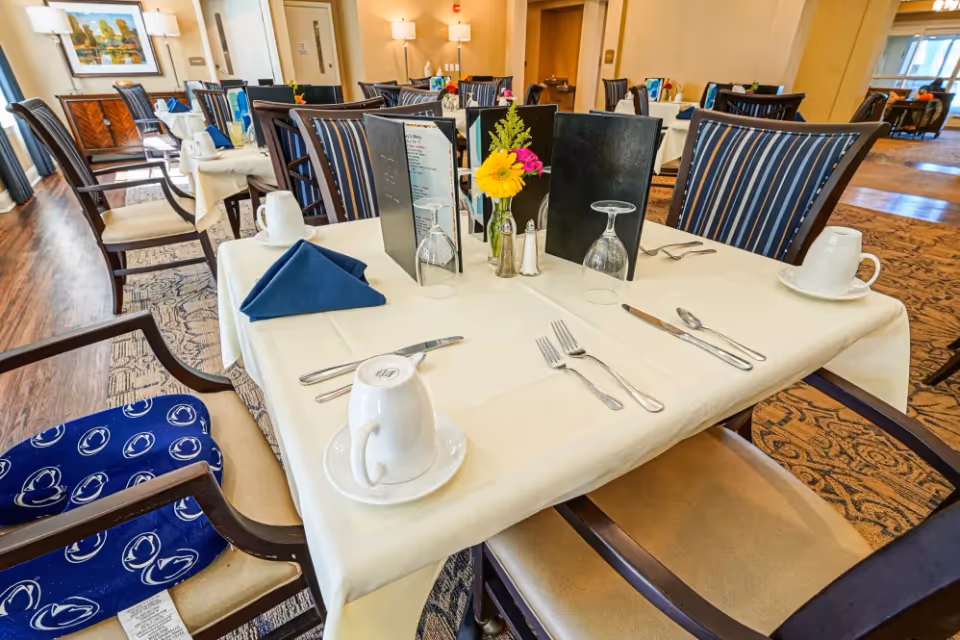 A neatly set dining table in a senior living facility dining room with white tablecloth, blue napkins, menus, glassware, silverware, coffee cups, and a small vase with yellow and pink flowers. The room has wooden floors, patterned carpet, and striped upholstered chairs.