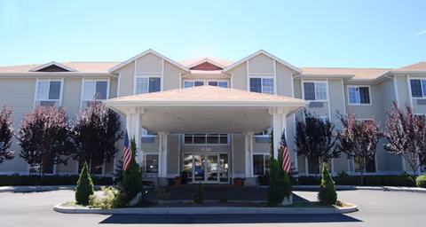 Front exterior view of a two-story senior living facility building with a covered entrance, multiple windows, and American flags on either side of the entrance. There are small trees and shrubs planted in front of the building under a clear blue sky.