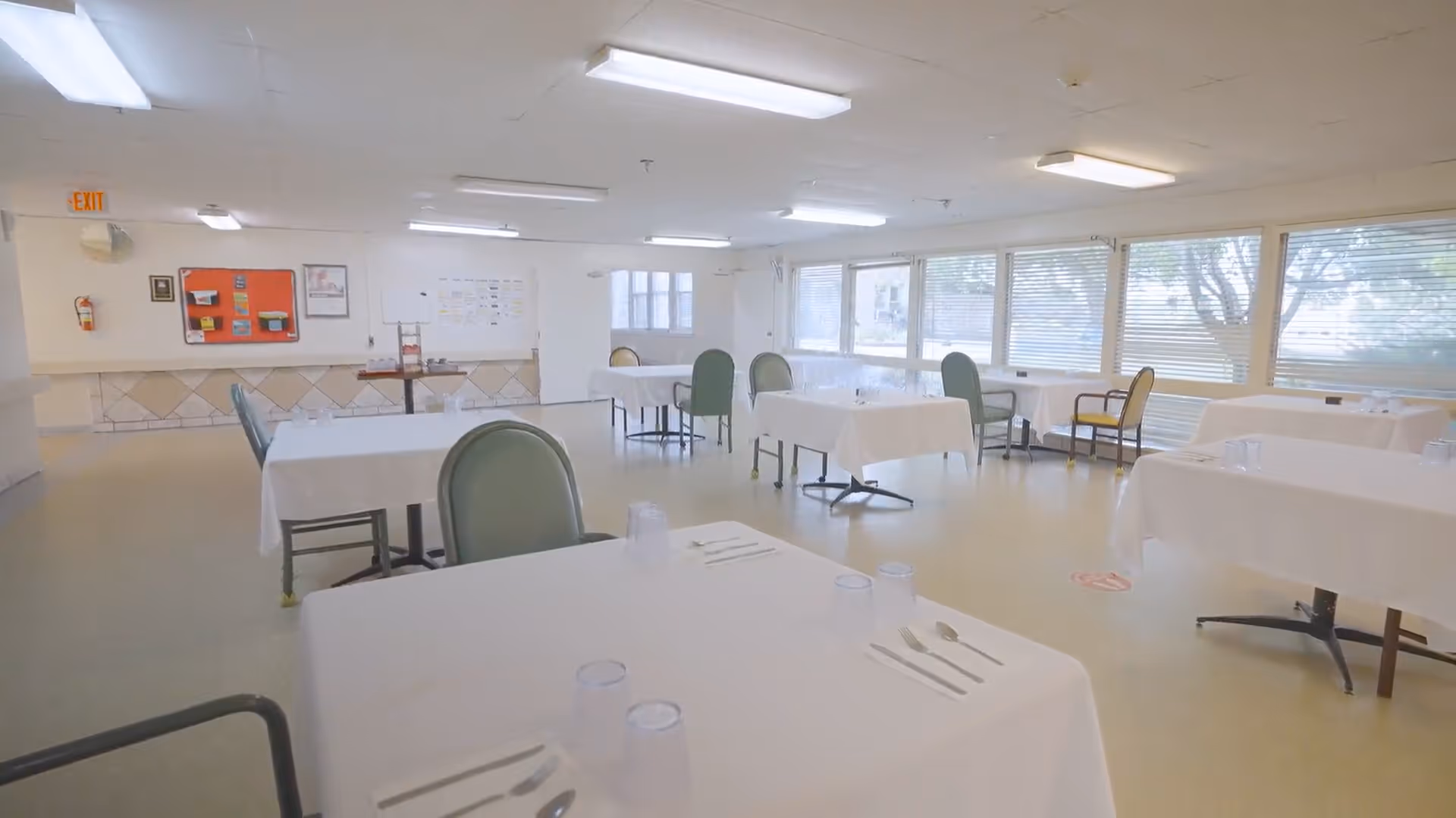 A bright dining room with several tables covered in white tablecloths, each set with utensils and clear plastic cups. The room has large windows with blinds, green and beige chairs, and fluorescent ceiling lights. A red bulletin board and an exit sign are visible on the far wall.