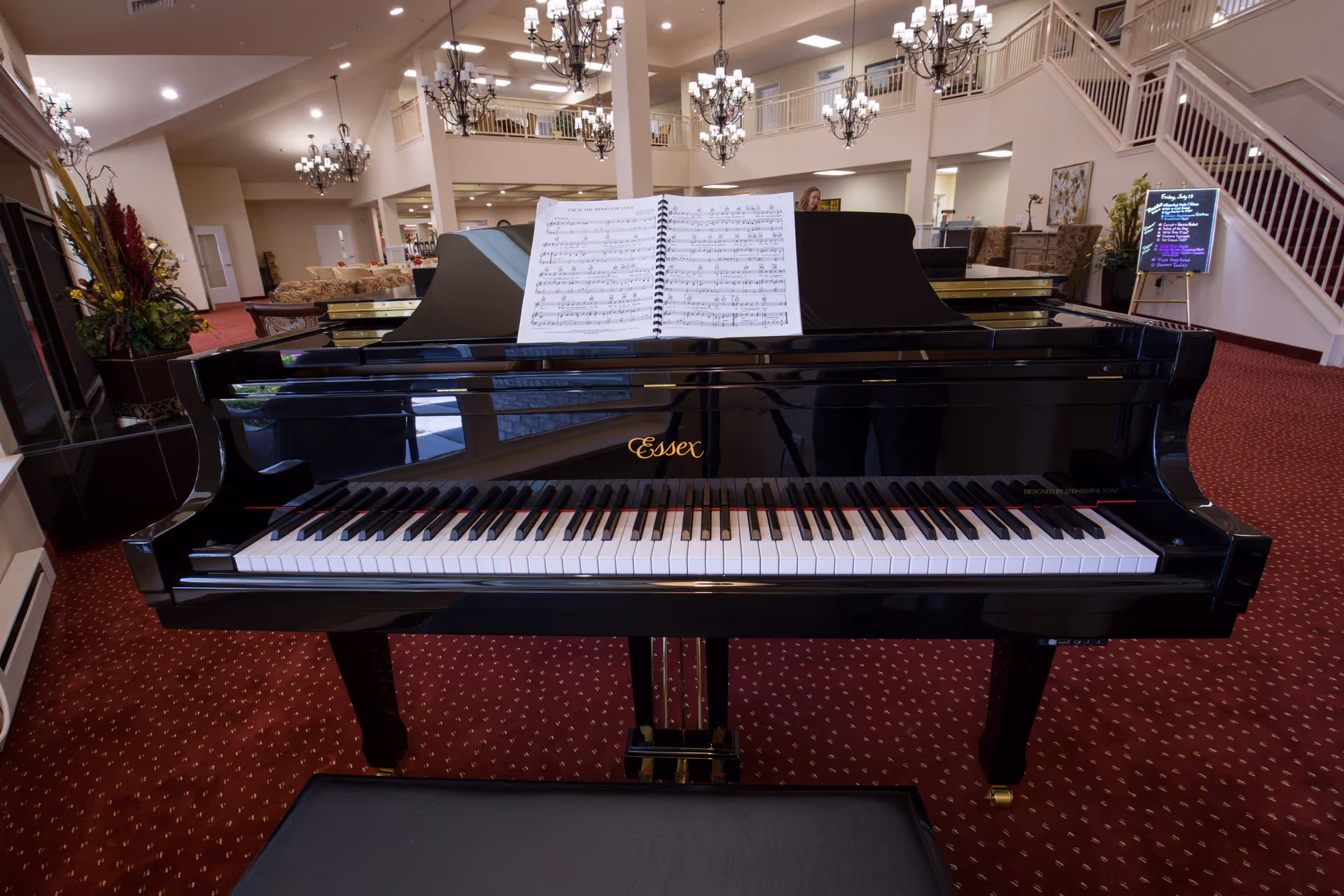 Black grand piano with sheet music in a senior living lobby featuring chandeliers, red carpet, and seating.