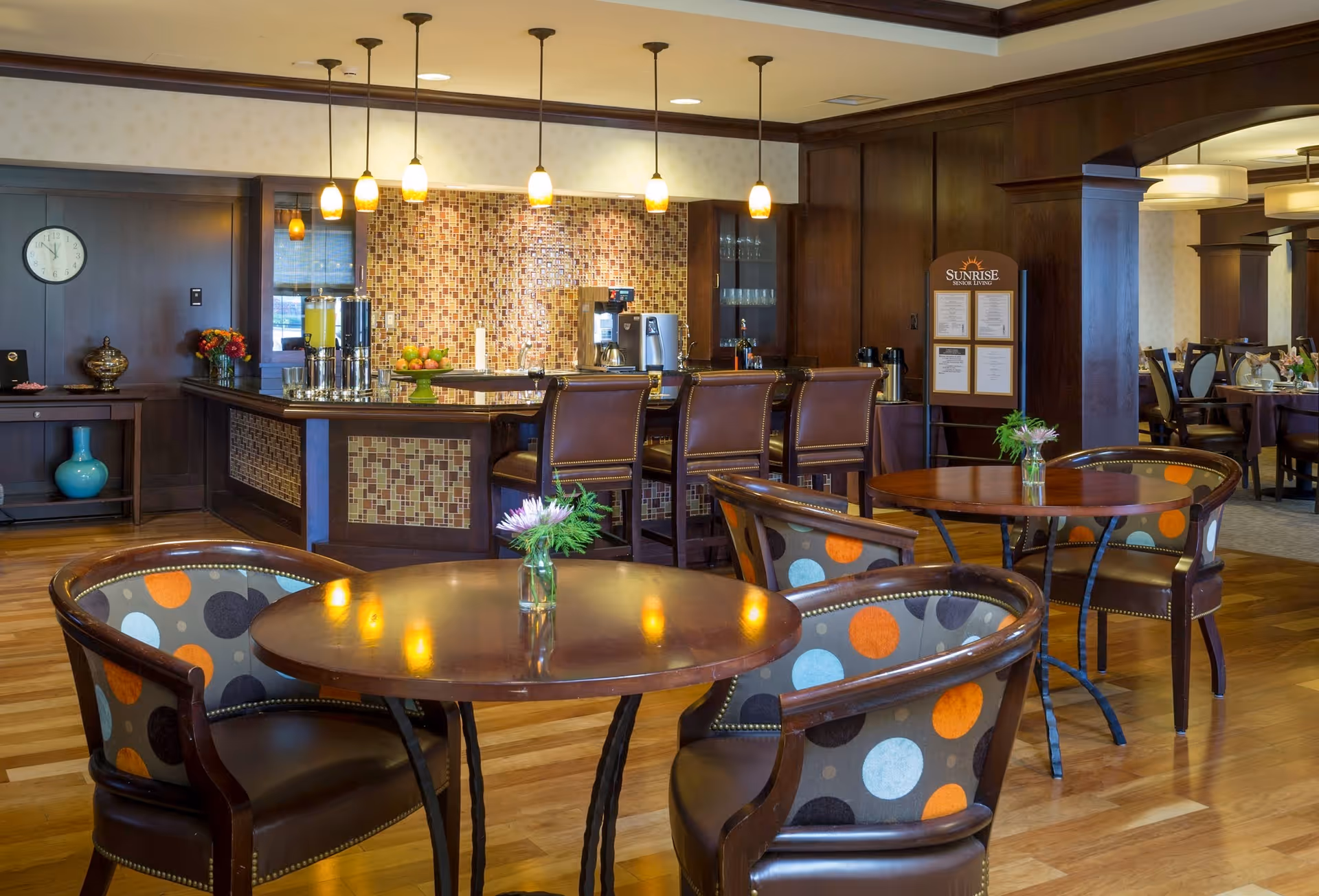 A cozy dining area in a senior living facility featuring round wooden tables with colorful polka dot upholstered chairs. In the background, there is a bar counter with high chairs, beverage dispensers, and a coffee machine. Warm pendant lights hang from the ceiling, and a menu board with the Sunrise logo is visible near the bar. The room has wooden flooring and dark wood paneling on the walls.