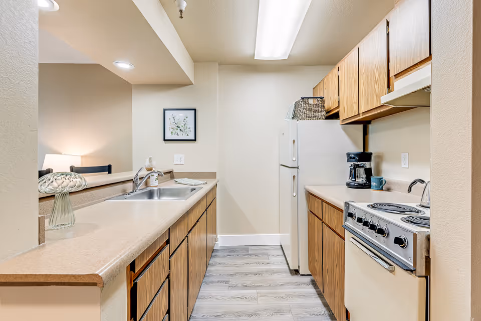 A compact kitchen with wooden cabinets, a white refrigerator, an electric stove, a coffee maker, and a sink. The kitchen has beige countertops and light-colored walls. There is a framed picture on the wall and a glass decorative item on the counter. The floor is covered with light gray wood-like flooring.