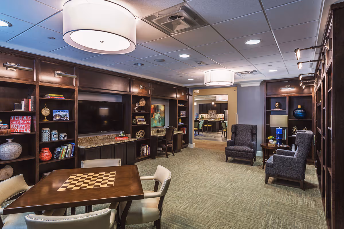 A well-lit community living room with dark wood built-in shelving and a TV, a chess table in the foreground, and upholstered armchairs.